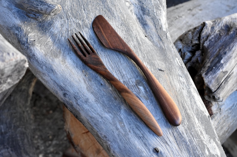 May include: A wooden fork and knife set made from dark brown walnut wood. The utensils are lying on a weathered gray wooden surface.