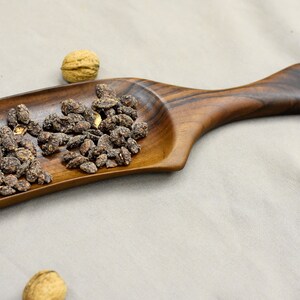 May include: A wooden serving tray with a long handle. The tray is filled with dark brown, roasted nuts.