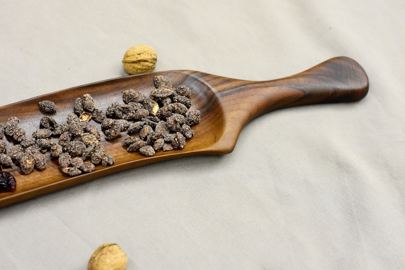 May include: A wooden serving tray with a long handle. The tray is filled with dark brown, roasted nuts.