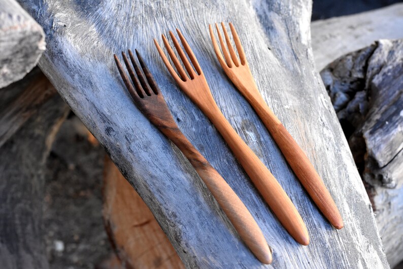 May include: Three wooden forks with different wood grain patterns. The forks are laid out on a piece of weathered wood.