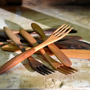 May include: A set of wooden utensils, including forks and spoons, made from different types of wood. The utensils are arranged in a pile on a light brown wooden surface.