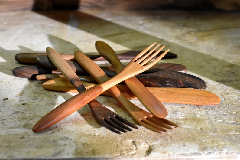 May include: A set of wooden utensils, including forks and spoons, made from different types of wood. The utensils are arranged in a pile on a light brown wooden surface.