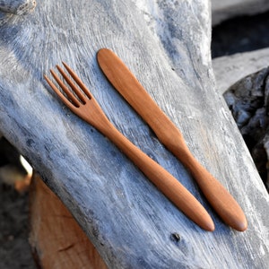 May include: A wooden fork and knife set on a weathered wooden surface. The utensils are made of natural wood and have a simple, rustic design.