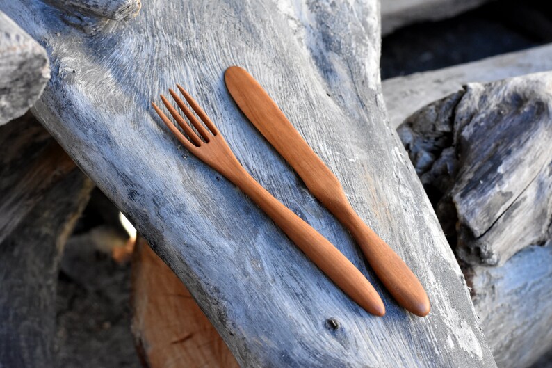 May include: A wooden fork and knife set on a weathered wooden surface. The utensils are made of natural wood and have a simple, rustic design.