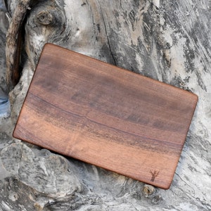 Puede incluir: Una tabla de cortar rectangular de madera de nogal. La tabla tiene un acabado de madera natural y un borde ligeramente redondeado. La tabla mide aproximadamente 30 cm de largo y 20 cm de ancho.