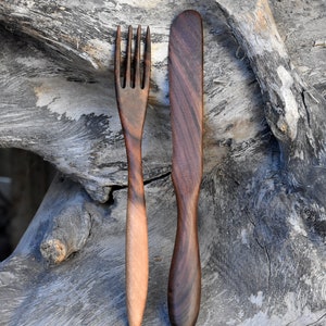 May include: A wooden fork and knife set, both with dark brown handles and natural wood grain. The utensils are placed on a weathered gray wooden surface.