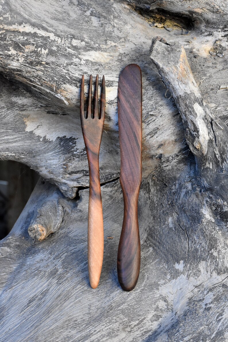 May include: A wooden fork and knife set, both with dark brown handles and natural wood grain. The utensils are placed on a weathered gray wooden surface.