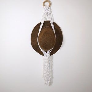 May include: White macrame hat holder with a brown hat hanging from it. The holder is attached to a white wall with a wooden ring at the top.