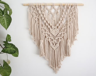 May include: Beige macrame wall hanging with intricate knotted patterns and fringe. The textile art piece is suspended from a natural wooden dowel. The design features geometric shapes and cascading strands, creating a bohemian aesthetic.