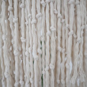 May include: A close-up of a white macrame wall hanging with a fringe of knotted cords.