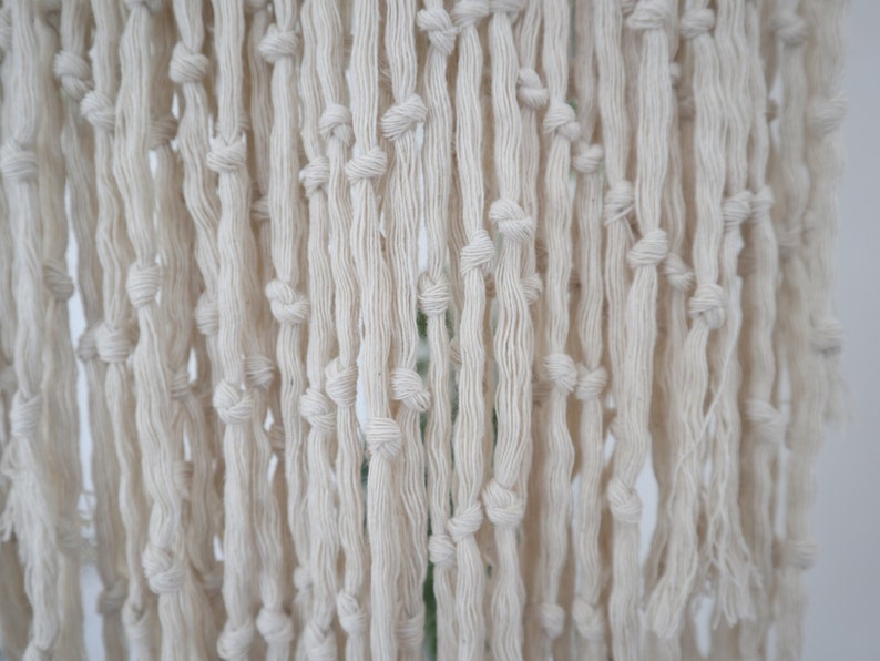 May include: A close-up of a white macrame wall hanging with a fringe of knotted cords.