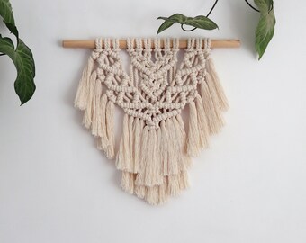 May include: A beige macrame wall hanging with a geometric pattern and fringe. The hanging is attached to a wooden dowel.