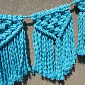 May include: A blue macrame banner with fringe. The banner is made of cotton rope and has a geometric pattern.