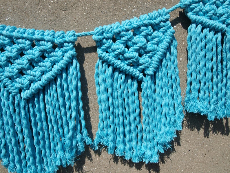 May include: A blue macrame banner with fringe. The banner is made of cotton rope and has a geometric pattern.