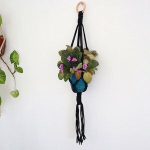 Può includere: Un portavasi in macramè nero con un anello di legno in cima, che tiene un vaso blu con una pianta verde e fiori rosa.