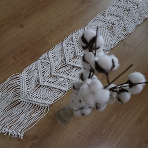 May include: White macrame table runner with a geometric pattern and fringe. The runner is laying on a wooden surface with a vase of white cotton flowers.