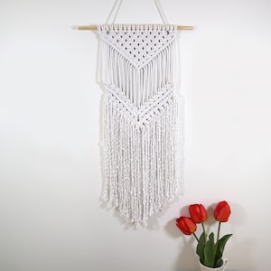 May include: White macrame wall hanging with a geometric pattern and fringe. The hanging is attached to a wooden dowel rod.
