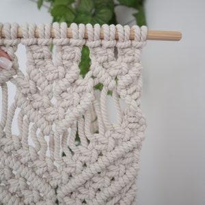 May include: A white macrame wall hanging with a geometric pattern. The hanging is attached to a wooden dowel rod.
