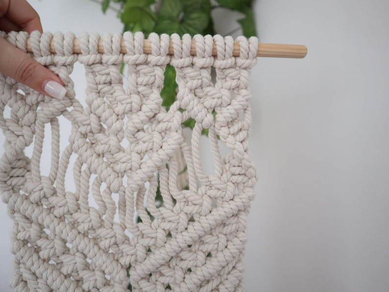 May include: A white macrame wall hanging with a geometric pattern. The hanging is attached to a wooden dowel rod.