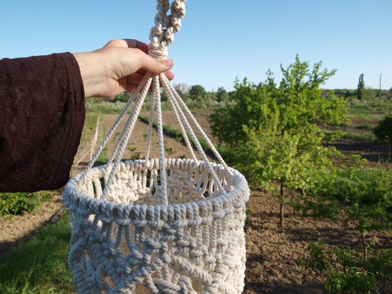 May include: A white macrame plant hanger with a round base and a knotted top. The hanger is made of thick cotton rope and has a delicate pattern.