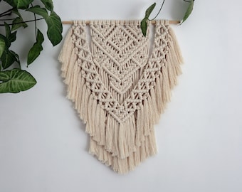 May include: A beige macrame wall hanging with a geometric pattern and fringe. The hanging is made of natural cotton cord and is attached to a wooden dowel.