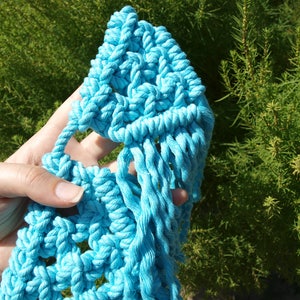 May include: A hand holding a blue macrame wall hanging with a fringe detail. The macrame is made with thick rope and has a geometric pattern.