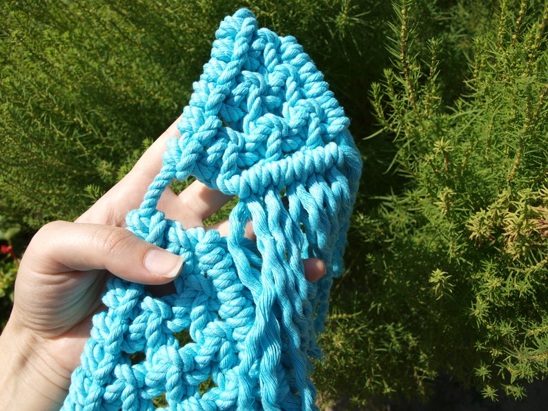 May include: A hand holding a blue macrame wall hanging with a fringe detail. The macrame is made with thick rope and has a geometric pattern.
