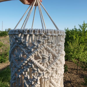 May include: A white macrame hanging lampshade with a geometric pattern and fringe. The lampshade is hanging from a rope and is in front of a green background.