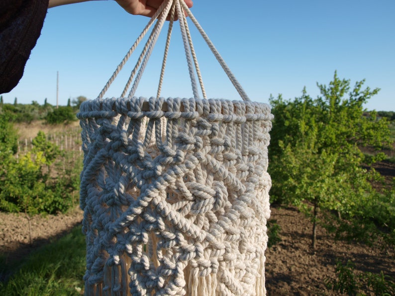 May include: A white macrame hanging lampshade with a geometric pattern and fringe. The lampshade is hanging from a rope and is in front of a green background.