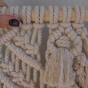 May include: A close-up of a macrame wall hanging with a wooden dowel at the top. The macrame is made with natural colored cotton cord and features a large tassel at the bottom.