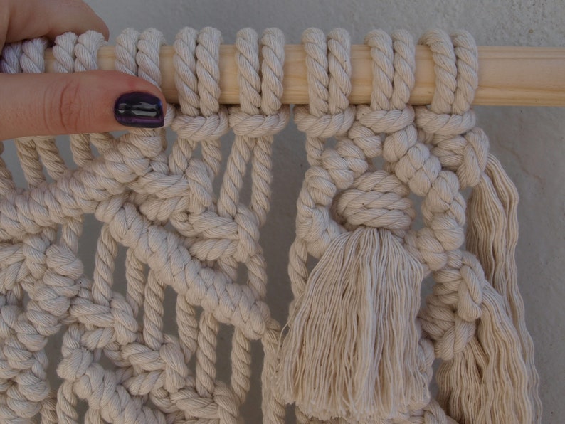 May include: A close-up of a macrame wall hanging with a wooden dowel at the top. The macrame is made with natural colored cotton cord and features a large tassel at the bottom.
