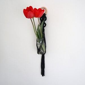 May include: A black macrame plant hanger with a wooden ring at the top, holding a glass vase with red tulips.