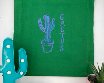 May include: A green tote bag with a purple and blue cactus design and the word "CACTUS" printed vertically. The bag has two green handles and is made of a sturdy fabric. The design features a potted cactus illustration.