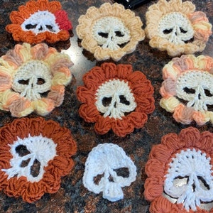 May include: A collection of crocheted skull-shaped flowers in various colors, including orange, yellow, white, and brown. The flowers are arranged on a dark surface.