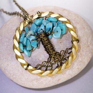 Op de afbeelding: Een handgemaakte hangerketting met een levensboomontwerp. De hanger heeft een goudkleurig gevlochten frame, een draadboomstam en turquoise gekleurde stenen bladeren. De ketting wordt gepresenteerd op een lichtgekleurde steen.
