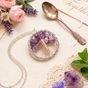 Pode incluir: Um colar de prata com um pingente em forma de árvore. O pingente é feito de fio de prata torcido e preenchido com pedras de ametista roxas. O colar é exibido com flores e uma colher vintage.