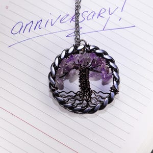 May include: A silver-toned pendant necklace featuring a tree of life design. The pendant has a circular frame with a braided edge, filled with purple gemstones and a detailed wire tree. The word "anniversary!" is written in purple ink on the lined paper background.