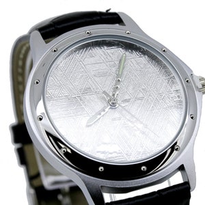Pode incluir: Um rel&oacute;gio de pulso prateado com uma pulseira de couro preto. O mostrador do rel&oacute;gio apresenta um design texturizado &uacute;nico que se assemelha a um meteorito.