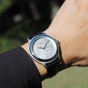 Pode incluir: Um rel&oacute;gio de pulso prateado com uma pulseira de couro preta. O mostrador do rel&oacute;gio &eacute; feito de um meteorito com um padr&atilde;o &uacute;nico.