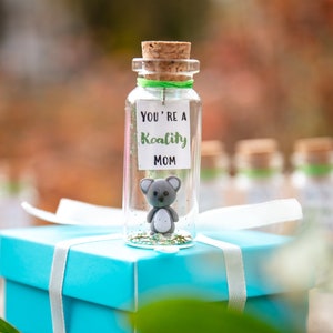 K&ouml;nnte beinhalten: Eine kleine Glasflasche mit einem Korkverschluss, gef&uuml;llt mit Glitzer und einer kleinen grauen Koala-Figur. Die Flasche hat ein wei&szlig;es Etikett mit dem Text "You're a Koality Mom". Die Flasche steht auf einer hellblauen Geschenkbox mit einem wei&szlig;en Band.