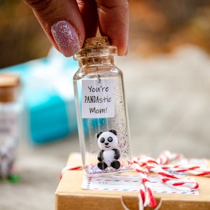 May include: A small glass bottle with a cork stopper contains a small panda figurine and purple glitter. The bottle has a white label with the text "You're PANDASTIC Mom!" and is sitting on a brown cardboard box with a red and white string tied around it. The box has a "This Side Up" label.