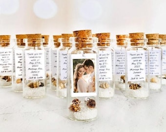 May include: A collection of clear glass bottles with cork stoppers, each containing white sand and small seashells. A rectangular tag is attached to each bottle, featuring a photograph of a couple and text reading: Thank you for celebrating with us May 27th, 2023 Montego Bay, Jamaica Love, Jeff & Leslie.