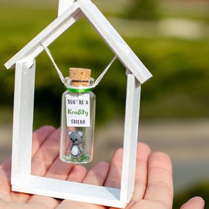 K&ouml;nnte beinhalten: Ein kleines wei&szlig;es Holzhaus mit einer Glasflasche, die im Inneren h&auml;ngt. Die Flasche enth&auml;lt eine kleine graue Koala-Figur und gr&uuml;nen Glitzer. Die Flasche hat einen Korkverschluss und ein wei&szlig;es Etikett mit der Aufschrift "You're a Koality Friend".