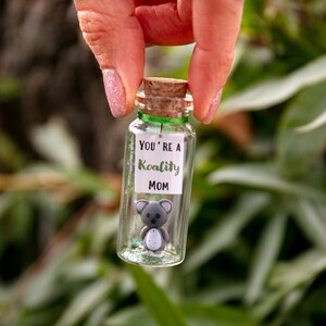 K&ouml;nnte beinhalten: Ein kleines Glas mit einem Korkverschluss und einem gr&uuml;nen Band um den oberen Rand. Das Glas enth&auml;lt Glitzer und eine kleine graue Koala-Figur. Ein wei&szlig;es Etikett auf der Vorderseite des Glases lautet "You're a Koality Mom".