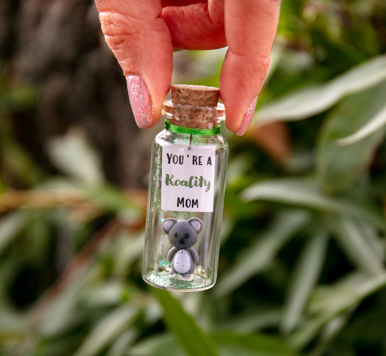 K&ouml;nnte beinhalten: Ein kleines Glas mit einem Korkverschluss und einem gr&uuml;nen Band um den oberen Rand. Das Glas enth&auml;lt Glitzer und eine kleine graue Koala-Figur. Ein wei&szlig;es Etikett auf der Vorderseite des Glases lautet "You're a Koality Mom".