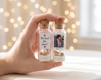 May include: Two clear glass bottles with cork stoppers, each containing white sand, small seashells, and a white pearl. One bottle features a tag reading We tied the knot! with red hearts, while the other displays a photograph of a couple and the text Keith & Rene 7/8/26.