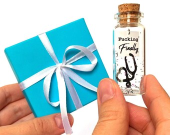May include: A bright turquoise square gift box tied with a white satin ribbon, accompanied by a small clear glass bottle with a cork stopper. Inside the bottle is a white paper tag with the text Fucking Finally and a black stethoscope charm.