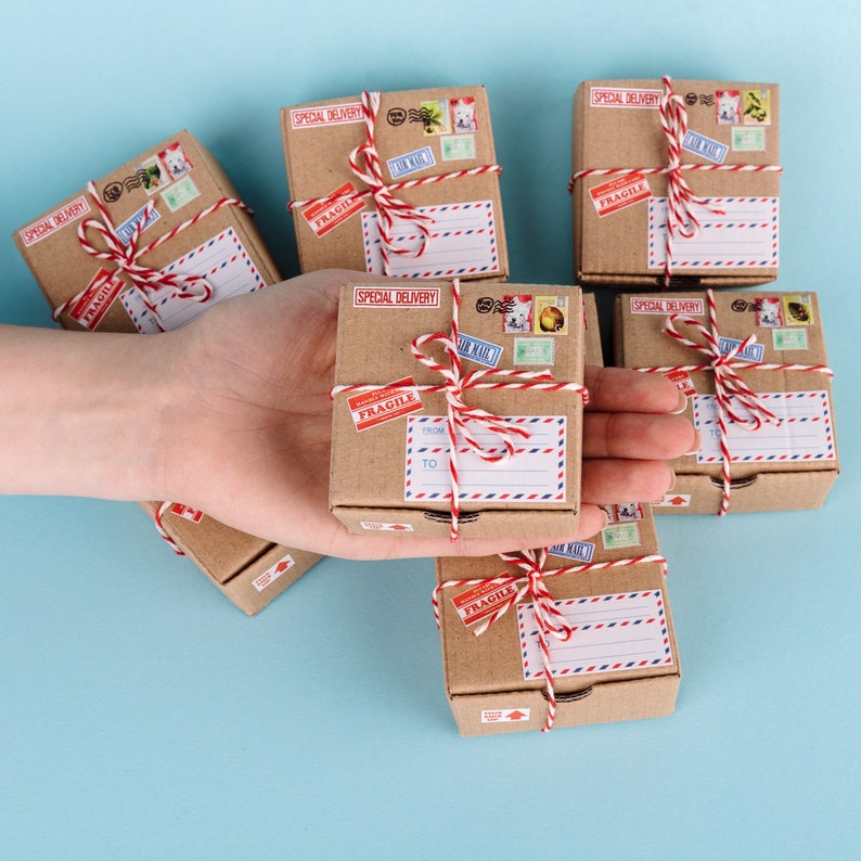K&ouml;nnte beinhalten: Sechs kleine braune Kartons, die mit rotem und wei&szlig;em Garn gebunden sind. Jede Schachtel hat ein "Special Delivery" und "Fragile" Etikett und ist mit Vintage-Briefmarken verziert.