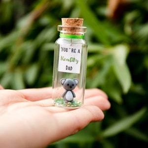 K&ouml;nnte beinhalten: Ein kleines Glasgef&auml;&szlig; mit einem Korkverschluss enth&auml;lt eine graue Koala-Figur und gr&uuml;nen Glitter. Ein wei&szlig;es Etikett auf dem Glas lautet "You're a Koality Dad."