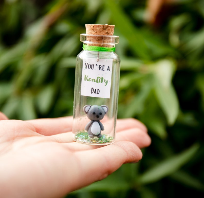 K&ouml;nnte beinhalten: Ein kleines Glasgef&auml;&szlig; mit einem Korkverschluss enth&auml;lt eine graue Koala-Figur und gr&uuml;nen Glitter. Ein wei&szlig;es Etikett auf dem Glas lautet "You're a Koality Dad."